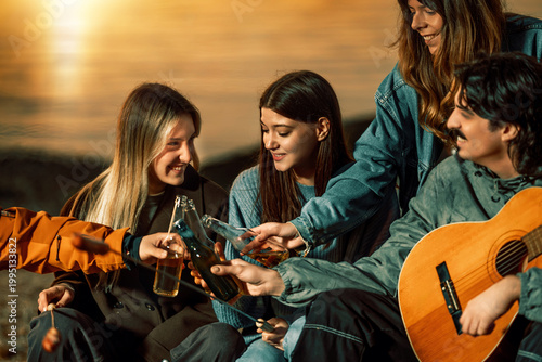 Friends share drinks and music by the water during sunset near a beach on a warm evening