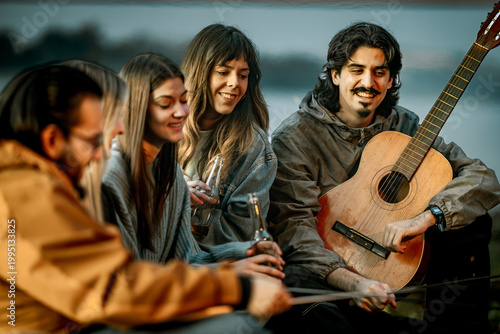 Group of friends enjoying time together near water while playing music and having drinks during sunset