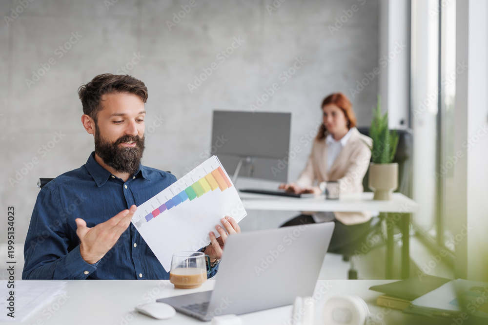 Fototapeta premium Man presents chart during meeting while woman works on laptop in modern office setting