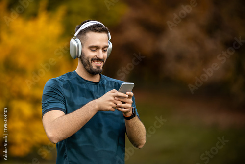 Man listens to music on phone while standing in park during autumn season