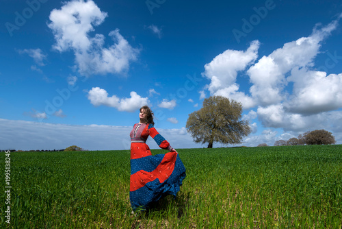Young woman in a red dress in a green field with sky and clouds 