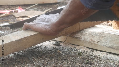 A carpenter is cutting a block of wood using a manual handsaw in an outdoor work area.