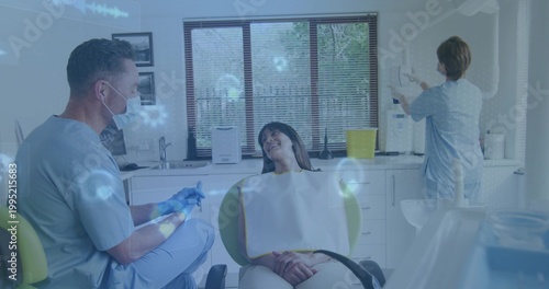 Treating dentist in blue scrubs wearing mask and gloves attending patient in operatory, with tray