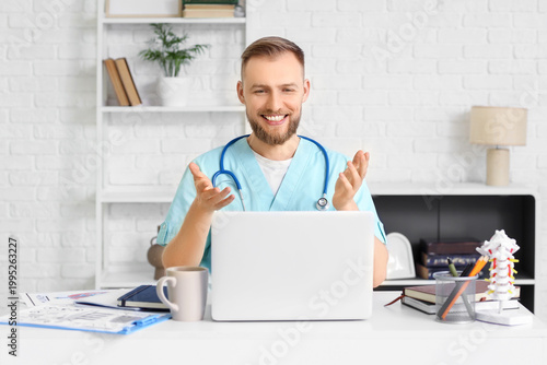 Male medical intern video chatting with laptop at table in clinic