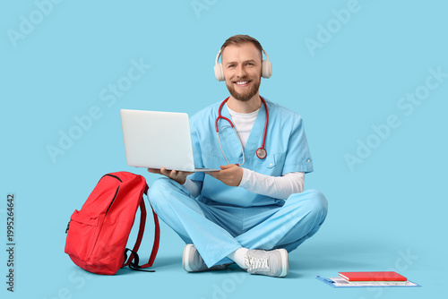 Handsome medical intern in headphones with backpack using laptop on blue back...