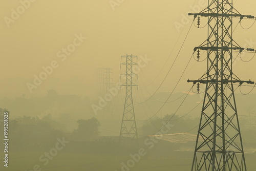 High voltage power pylons in foggy sunrise scene, energy infrastructure across rural landscape with warm light, haze and layered silhouettes in countryside