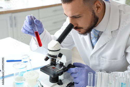 Male scientist with blood sample looking through microscope at table in labor...