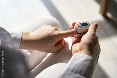 Person Checking Oxygen Saturation With Fingertip Pulse Oximeter While Holding Finger Over Device While Seated In Bright Indoor Setting With Sunlight On Lap And White Clothing.