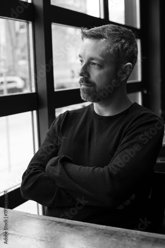 Black and white portrait of a man in his 40s seated by a restaurant window in Ottawa, Canada. Thoughtful side profile with soft natural light, casual sweater, and calm modern atmosphere indoors.