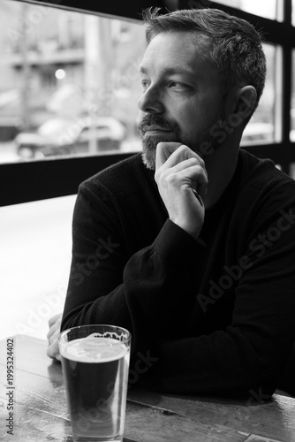 Black and white portrait of a man in his 40s seated by a restaurant window in Ottawa, Canada. Thoughtful side profile with soft natural light, casual sweater, and calm modern atmosphere indoors.