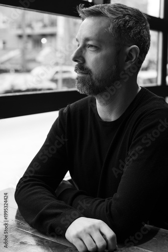 Black and white portrait of a man in his 40s seated by a restaurant window in Ottawa, Canada. Thoughtful side profile with soft natural light, casual sweater, and calm modern atmosphere indoors.