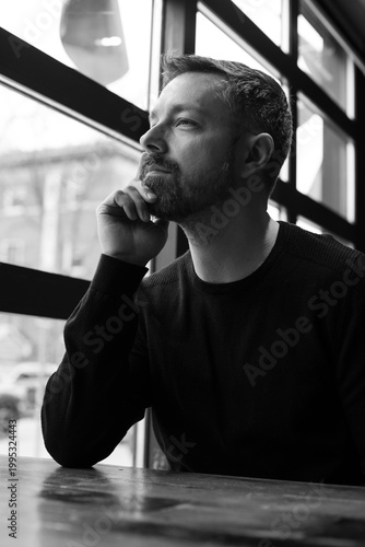 Black and white portrait of a man in his 40s seated by a restaurant window in Ottawa, Canada. Thoughtful side profile with soft natural light, casual sweater, and calm modern atmosphere indoors.