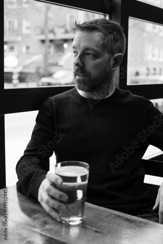Black and white portrait of a man in his 40s seated by a restaurant window in Ottawa, Canada. Thoughtful side profile with soft natural light, casual sweater, and calm modern atmosphere indoors.