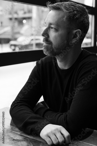 Black and white portrait of a man in his 40s seated by a restaurant window in Ottawa, Canada. Thoughtful side profile with soft natural light, casual sweater, and calm modern atmosphere indoors.