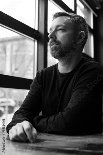 Black and white portrait of a man in his 40s seated by a restaurant window in Ottawa, Canada. Thoughtful side profile with soft natural light, casual sweater, and calm modern atmosphere indoors.