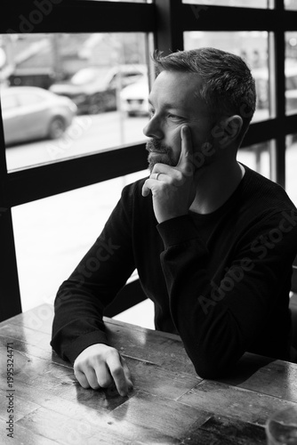 Black and white portrait of a man in his 40s seated by a restaurant window in Ottawa, Canada. Thoughtful side profile with soft natural light, casual sweater, and calm modern atmosphere indoors.