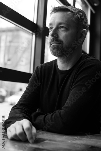 Black and white portrait of a man in his 40s seated by a restaurant window in Ottawa, Canada. Thoughtful side profile with soft natural light, casual sweater, and calm modern atmosphere indoors.