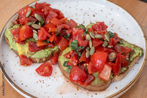 Two slices of whole-grain toast generously topped with mashed avocado, fresh diced red tomatoes, basil, and crunchy pumpkin seeds on a speckled white plate.