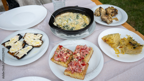 A diverse spread of authentic Albanian dishes, including bruschetta, eggplant, roast chicken, pastice, and a creamy gratin, adorns a rustic dining table, in Theth, Albania.