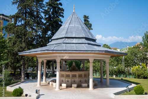 Exterior view of a traditional wudu fountain with a layered conical roof, white columns, and intricate details, set within a manicured green area under a bright blue.