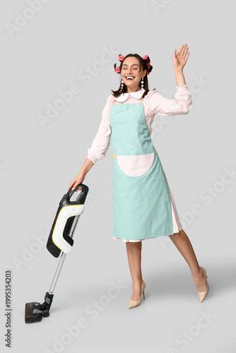 Obraz na płótnie Portrait of happy young housewife in apron with vacuum cleaner on grey backgroun