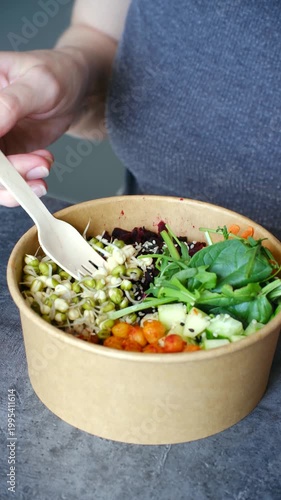 Woman hands eating fresh buddha bowl with wooden fork, healthy meal with mung bean sprouts, chickpeas, spinach, beetroot, cucumber in craft paper container for takeout or delivery. Vertical shot