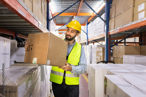 Фотография Male warehouse worker wearing safety vest and hardhat, holding cardboard box standing in industrial storage facility