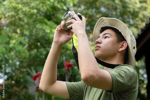 A young man in a boonie hat is taking a photo with his camera, looking focused and passionate about photography in a lush, outdoor environment.