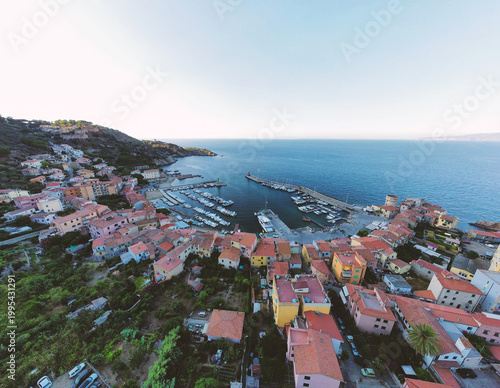 Aerial view of the harbor of Giglio Porto featuring colorful houses, docked boats, and a stone pier along the coastline in Giglio Porto, Tuscany, Italy.