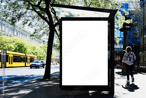 blank lightbox and glass bus shelter at bus stop. soft urban street background. white ad poster. display panel. empty outdoor ad space. mockup base. yellow tram, car and person passing by