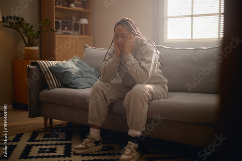 Young adult woman sitting with electronic ankle monitor, holding head on sofa during house arrest. Useful for legal restriction, criminal justice, surveillance, confinement themes