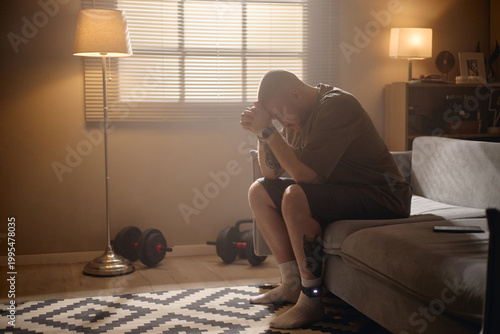 Obraz na płótnie Young adult man sitting with electronic ankle monitor, bowing head and clasping hands at home, representing house arrest and legal restriction