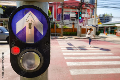 Close-up of a pedestrin crossing button with an upward arrow symbol at a crosswalk in an urban area. The background shows a blurred city street with a person walking across the zebra crossing.