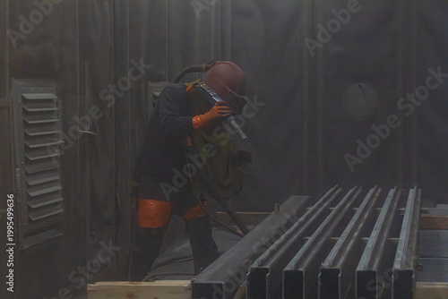 Fotografie Artisan en combinaison de protection utilisant une lance de sablage à haute pression pour décaper des profilés métalliques dans une cabine industrielle