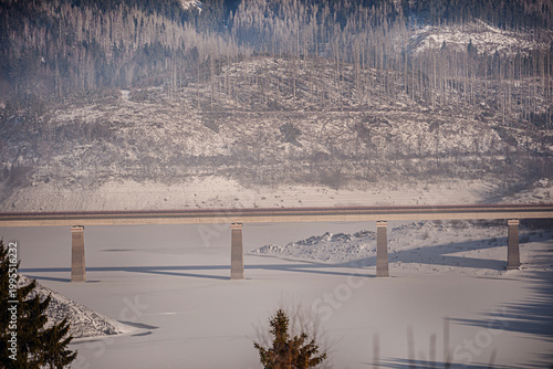Modern concrete bridge crossing a frozen snow covered reservoir in misty winter mountains