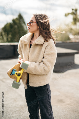 Young woman in a fluffy jacket holding a skateboard on a sunny day