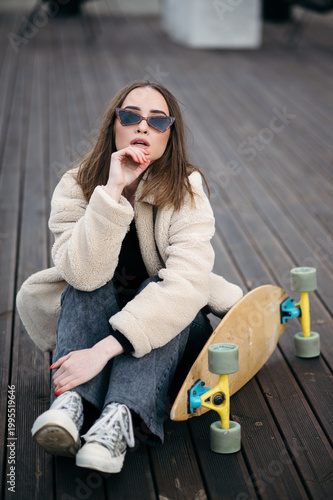 Young woman in stylish outfit with skateboard sitting on wooden deck