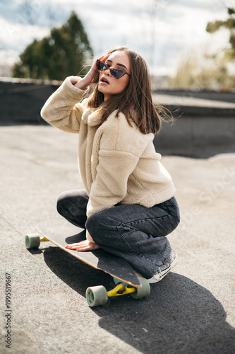 Young brunette squatting near skateboard on rooftop