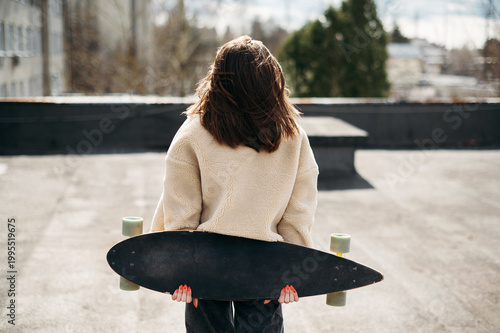 Back view of young brunette posing with skateborad on roof