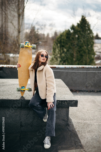 Young woman in stylish wear posing on roof with skateboard
