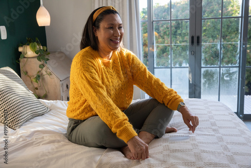 Quadro su tela Mature woman sitting cross-legged on bed at home, wearing yellow sweater, touchi