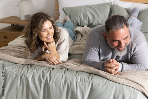 Romantic couple lying on bed in bedroom, propping on forearms, smiling with sage-green sheets
