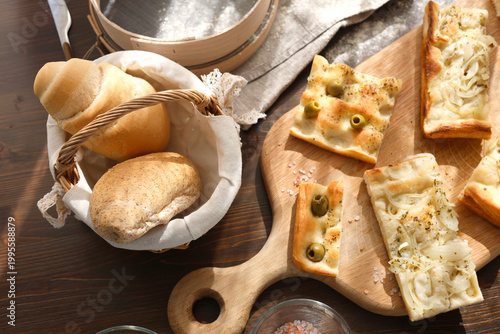 Assorted italian bread with focaccia and olives on wooden board. Top view of fresh bakery products including rolls in basket and sliced flatbread with herbs. Professional food photography shows