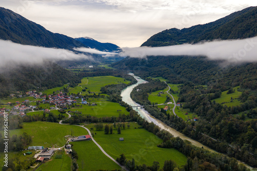 Aerial view of the Soca River winding through a lush green valley with the village of Srpenica nestled among forested mountains and low clouds Srpenica, Tolmin, Slovenia.
