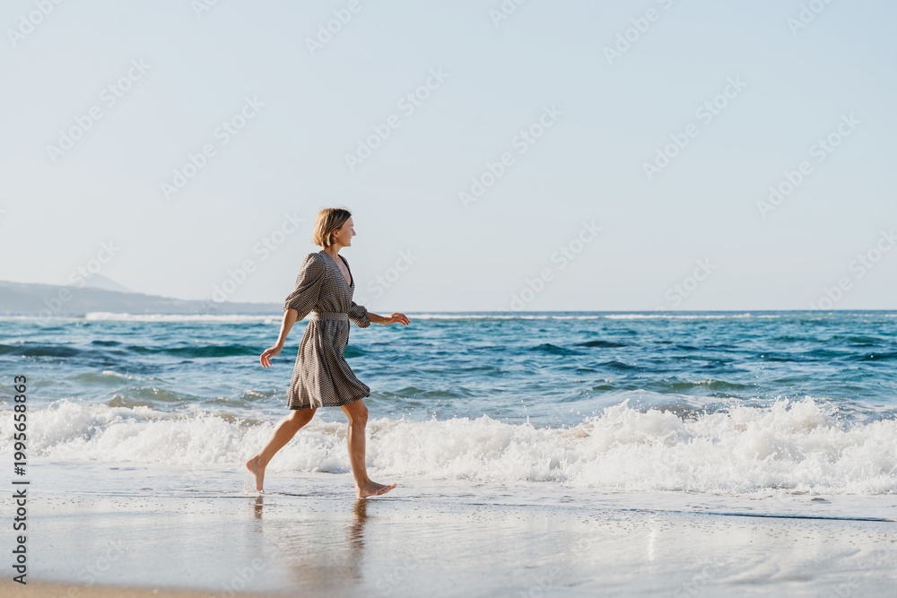 Obraz premium Happy woman runs and jumps by the ocean. Concept freedom and carefree vacation. Beach Holidays. Carefree woman splashing water having fun on the beach. Young girl enjoying fresh air on ocean shore.