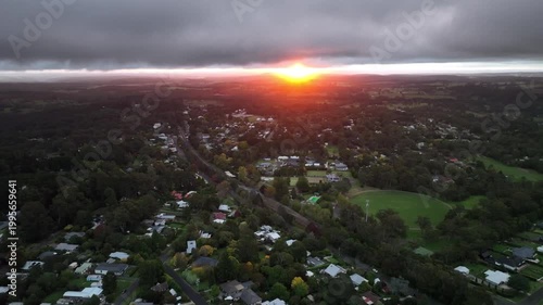 Dramatic low-altitude drone footage of a fiery orange and red sunrise over a green paddock in Bundanoon, Southern Highlands, New South Wales, Australia. Stormy clouds above an intense glowing horizon.