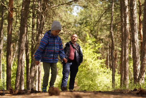 A young boy in a blue striped jacket and beanie walks along a sun-dappled dirt path through a pine forest, with a man following behind him, capturing a moment of family exploration.