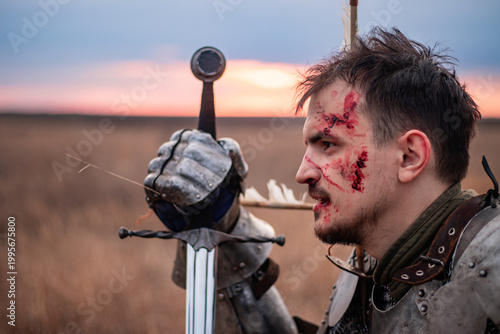 Close up portrait knight with a sword in hand, wounded by arrows sits in the field on the dry grass after a battle, background of sunset. defeated medieval warrior kneels in a field with a bloody face
