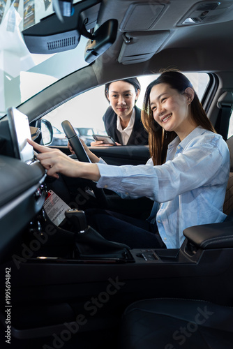 Asian female car salesperson explaining interior features to a happy young Asian woman customer sitting in a new car at the dealership showroom. Vertical Shot
