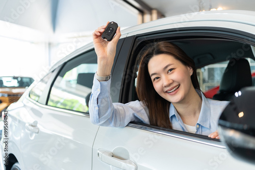 Happy young Asian woman smiling inside a new white car at an auto dealership showroom, proudly showing her new car key. Concept of buying a new vehicle.